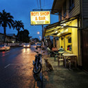 There's something profoundly grounding about the simple architecture of community gathering spaces, especially on a quiet Tuesday evening when the rain has just passed through. I came across this roti shop tonight on my way back from a community consultation meeting in the Rainville district, and I had to pause. The warm amber glow spilling onto the wet pavement, the hand-painted signage, the casual guardian dog at the entrance—these are the elements that make a neighborhood feel truly lived-in and loved.
As urban planners and architects, we often get caught up in grand visions of green corridors, sustainable infrastructure, and large-scale public interventions. And while those projects are undeniably important—they're the work that keeps me up at night, sketching and recalculating—there's equal value in recognizing and preserving these modest, authentic spaces that already serve as social anchors in our communities. This roti shop isn't just a place to eat; it's a node in the social network of this neighborhood, a point of connection where people gather after work, where conversations happen naturally, where the rhythm of daily life is maintained.
The reflection of light on wet asphalt creates an almost painterly quality here, doesn't it? It reminds me why I returned to Paramaribo after my studies in Delft. The Netherlands taught me technical excellence and gave me exposure to cutting-edge sustainable design principles, but Suriname—this is where the heart of community-centered architecture truly reveals itself. It's in these unplanned moments of beauty, in the organic way spaces evolve to meet people's needs, in the resilience of small businesses that weather both literal and metaphorical storms.
I've been thinking a lot lately about how our urban greening strategy can support rather than displace these existing community touchstones. How do we bring more green infrastructure, better pedestrian connectivity, and climate-adaptive design to neighborhoods while ensuring that places like this—with their character, their affordability, their essential role in the social fabric—remain viable and valued? It's one of those complex challenges that requires us to listen more than we prescribe, to design with humility and cultural sensitivity.
Tonight, looking at this simple scene, I'm reminded that sustainable urban development isn't just about environmental metrics or aesthetic transformation. It's about understanding what makes a place feel like home to the people who live there, and building from that foundation of respect and recognition.