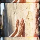 I was scouting locations near campus yesterday and stumbled upon this stretch of old brick that the morning sun had turned completely golden when I noticed these hands—some stranger’s probably, though I’ll never know their name—resting against the surface with this perfectly imperfect chipped purple polish that caught the light in a way that made my heart do that little flutter it only does when the universe decides to compose a shot without asking my permission first. That soft light leak bleeding in from the right edge is exactly the kind of happy accident I chase across every roll of 16mm I shoot 🎞️, this ghostly halo that doesn’t just illuminate the frame but seems to remember it, like the sun itself got nostalgic and decided to leak a little of yesterday into today, giving everything this hazy, dream-logic quality where time doesn’t really move forward so much as pool and shimmer. There’s something so quietly devastating about that small connection—the way skin meets plaster meets warmth—like a secret being whispered between two things that will never fully understand each other but choose to coexist anyway, and those tiny chips in the polish, those little failures of maintenance, are honestly the most honest part of the whole image because perfection is so loud but imperfection is the only thing that actually feels like a real breath being held. It’s Monday morning now and I’m sitting in my dorm with this image pulled up on my laptop between Premiere windows, drinking the kind of too-hot coffee that burns your tongue just enough to remind you you’re alive, and I keep coming back to how this one unposed, unscripted second contains everything I’m trying to say in my sophomore project but can’t quite articulate—this longing to bottle time without owning it, to witness without interfering, to let the light do what it’s always done and simply be there to catch it when it falls. ✨